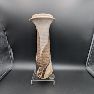 May include: A handmade ceramic vase with a flared top and a narrow base. The vase is light beige with brown and black abstract patterns. The vase is displayed on a clear acrylic stand. The vase is approximately 25 cm tall.