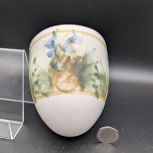 Könnte beinhalten: Eine weiße Keramiktasse mit einem bemalten Blumendesign in Blau, Grün und Braun. Die Tasse hat eine abgerundete Form und einen hellbraunen Rand. Eine Münze liegt neben der Tasse.