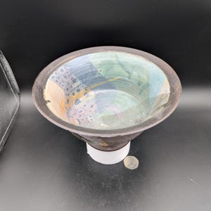 May include: A small, ceramic bowl with a dark brown rim and a colorful, abstract design inside. The bowl has a wide, shallow base and a slightly flared lip.