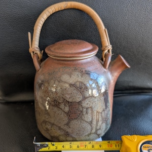 May include: A brown ceramic teapot with a woven handle and a decorative design. The teapot has a rounded body and a spout on the side.