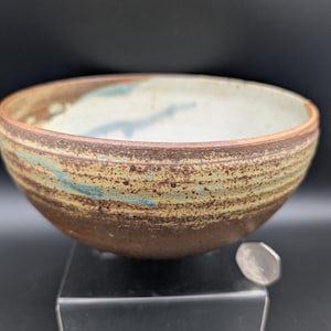 May include: A handmade ceramic bowl with a rustic, earthy aesthetic. The bowl features a brown exterior with bands of turquoise and tan, and a light interior. The bowl is likely 6-8 inches in diameter, perfect for serving or display.