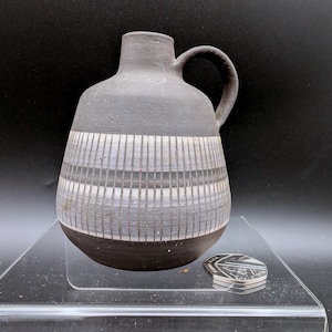 May include: A matte black ceramic jug with a narrow neck and a single handle. The body features a wide horizontal band of etched vertical white lines and grey stripes. A silver-coloured coin is placed next to the base for scale reference.