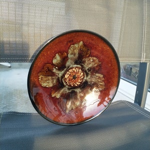 May include: A brown ceramic plate with a large, stylized flower design in shades of brown, green, and gold. The flower has a central circle with a smaller, concentric circle in the center.