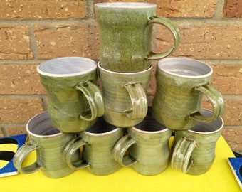 May include: A stack of eight green ceramic mugs with white interiors. The mugs are stacked in a pyramid shape, with the largest mug at the top and the smallest mugs at the bottom.