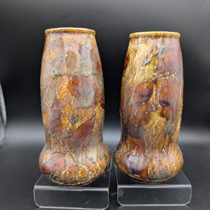May include: Two ceramic vases with a leaf pattern in shades of brown, tan, and blue. The vases have a bulbous base and a slightly flared rim. They are displayed on clear acrylic stands against a black background.