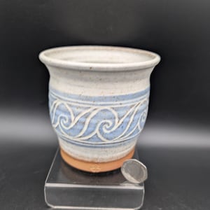 Può includere: Una tazza in ceramica maculata con bordo svasato e una fascia azzurra con un motivo a onde bianche. La base è in terracotta non smaltata con un piccolo marchio stampato. Una moneta è posizionata accanto per riferimento di scala.