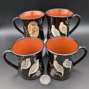 May include: Set of four black ceramic mugs with orange rims. Each mug features a different hand-painted design of birds and an owl on a branch.