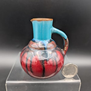 Peut inclure: Un petit pichet en céramique avec un corps arrondi et une anse. Le pichet présente une palette de couleurs vives, avec un dessus turquoise, une base rouge et des rayures verticales noires. Le pichet mesure environ 7.6 cm de haut.