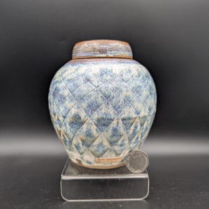 May include: A ceramic jar with a lid featuring a blue and white diamond-patterned glaze. The base of the jar has a label that reads ANVIL POTTERY Made in Wales. A silver-colored coin is placed next to the jar for scale on a clear stand.