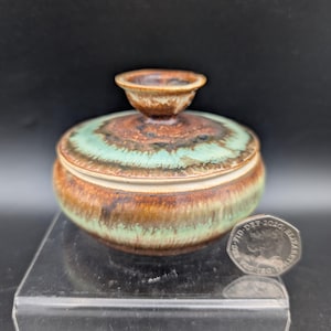 Op de afbeelding: Een kleine, ronde keramische pot met deksel, voorzien van een reactief glazuur met gemengde banden in aardebruin, crème en zeegroen. Het deksel heeft een opvallende, uitlopende knopgreep. Een Britse munt van twintig pence ligt ernaast.