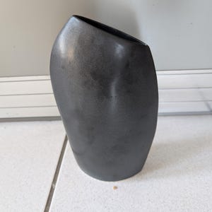 Op de afbeelding: Een donkergrijze, moderne vaas met een unieke, asymmetrische vorm. De vaas heeft een gladde, metallic afwerking en een schuine opening. Het is een decoratief item, geschikt voor het tentoonstellen van bloemen of als op zichzelf staand kunstwerk.