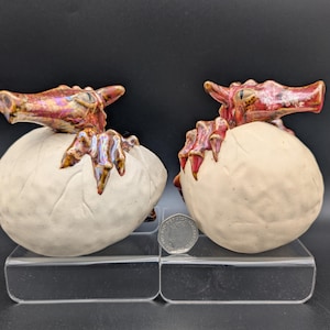 May include: Two ceramic dragon sculptures, each hatching from an egg. The dragons have red and gold scales, with detailed claws and faces. The eggs are off-white, and the sculptures are displayed on a clear acrylic surface.