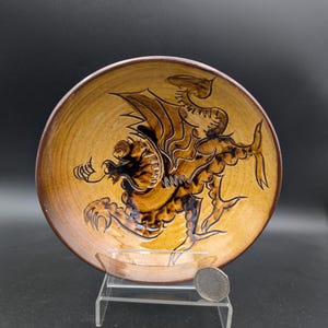 May include: A ceramic plate with a dragon design. The plate has a brown rim and a tan center with a detailed illustration of a dragon in shades of brown. The dragon is depicted with wings, claws, and a long tail. The plate is displayed on a clear stand.