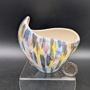 Könnte beinhalten: Eine kleine Keramikschale mit einer einzigartigen, asymmetrischen Form. Die Außenseite ist mit einem farbenfrohen, abstrakten Muster aus vertikalen Pinselstrichen in Gelb, Blau, Rosa und Schwarz verziert. Die Innenseite ist cremefarben. Die Schale steht auf einem transparenten Ständer.