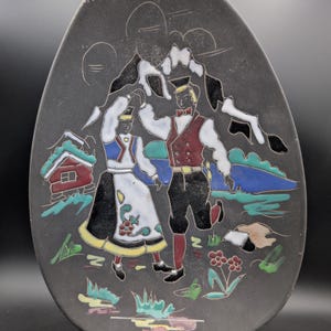 Puede incluir: Un plato decorativo negro de forma ovalada con un diseño de arte popular. La imagen muestra a una pareja con vestimenta tradicional bailando frente a montañas y una pequeña casa. El plato tiene un acabado brillante y probablemente sea de cerámica.