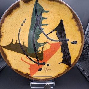 May include: A decorative ceramic plate with an abstract design. The plate features a yellow background with green, orange, and black shapes. The rim is a dark brown color. The plate is displayed on a clear stand.
