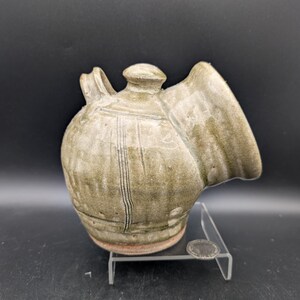 May include: A round, light green glazed ceramic vessel with a wide, angled opening on the right side, a small handle on the left, and a lid on top. A small silver coin is placed on the clear acrylic stand next to the base for scale.