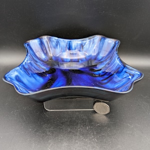 May include: A square glass bowl with a wavy, ruffled rim featuring a deep blue and black marbled pattern. The bowl is elevated on a clear acrylic stand, with a silver-coloured coin placed next to the base for scale, providing a visual reference.