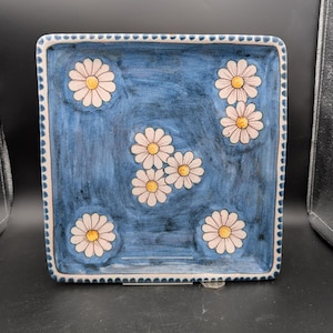 Peut inclure: Plat carré en céramique avec un fond bleu et un motif floral de marguerites blanches. Les marguerites ont des centres jaunes. Le bord est blanc avec un bord festonné. Le plat est probablement fait à la main.
