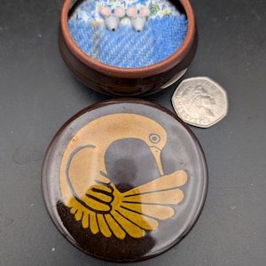 Può includere: Un portagioie in ceramica marrone con un coperchio con un disegno di uccello giallo e marrone. All'interno della scatola aperta, tre piccole figurine di topi grigi poggiano su un tessuto blu con motivi floreali e tweed. C'è una moneta da 50 pence accanto.