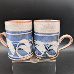 Può includere: Due tazze in ceramica con un motivo a foglie blu e bianco. Le tazze hanno un bordo e un manico marrone. Le tazze sono cilindriche e hanno un aspetto rustico e fatto a mano. Le tazze sono su un supporto trasparente su sfondo nero.