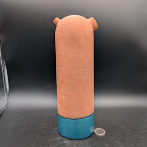 May include: A cylindrical, terracotta-colored ceramic object with two small ear-like protrusions at the top. The base is a band of teal blue. The object is standing upright.