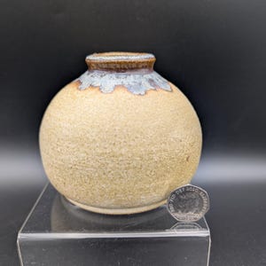 Può includere: Un piccolo vaso rotondo in ceramica con un corpo beige maculato e strutturato. Il vaso ha un bordo marrone con un bordo decorativo azzurro e bianco. Il vaso è esposto su un supporto in acrilico trasparente su sfondo nero.