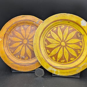 Op de afbeelding: Twee ronde keramische borden met een geel en bruin kleurenpalet. Elk bord toont een centraal zonnebloem- of bloemmotief met langwerpige bloemblaadjes. Een kleine munt is tussen de borden geplaatst als referentie voor de schaal.