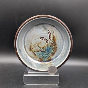 May include: A round ceramic bowl with a dark brown rim and a light blue, grey, and brown mottled interior glaze. The centre features a hand-painted design of green leaves and two stalks of wheat. A small silver coin is placed at the base for scale.