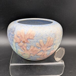 May include: A small, round ceramic bowl with a speckled light blue glaze and embossed, muted pink leaf-like patterns. The bowl is positioned next to a silver-coloured coin for scale, showcasing its compact size and textured, matte surface finish.