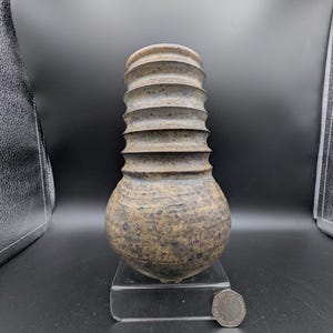 Könnte beinhalten: Eine handgefertigte Keramikvase mit rundem Boden und geriffeltem Hals. Die Vase hat eine gesprenkelte, erdige Farbpalette aus Braun, Beige und Grau. Die Vase wird auf einem transparenten Acrylständer ausgestellt.