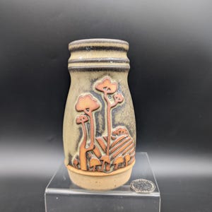 May include: A tan ceramic vase with a textured surface and a brown rim. The vase features a raised relief design of stylized orange mushrooms and trees. The vase is displayed on a clear acrylic stand.
