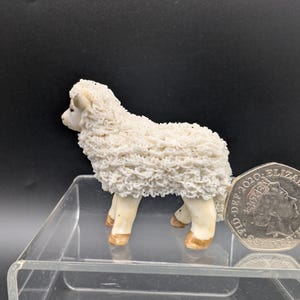 Könnte beinhalten: Kleine, cremefarbene Keramik-Schaf-Figur mit strukturiertem, wolligem Fell. Das Schaf hat hellbraune Hufe und ein detailliertes Gesicht. Die Figur wird auf einer transparenten Acrylfläche vor dunklem Hintergrund präsentiert.