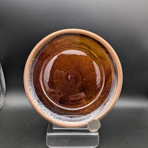 May include: A handmade ceramic plate with a glossy, dark brown center and a lighter brown rim. The plate has a textured surface and a slightly irregular shape, suggesting it is handcrafted. The plate is displayed on a clear stand against a dark background.