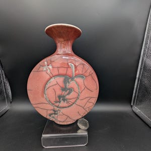 May include: A round, flat-bodied ceramic vase with a flared neck, featuring a dusty rose glaze with a prominent dark crackle pattern. A circular medallion with a dark, abstract calligraphic design is centered on the front, accompanied by small floral motifs.