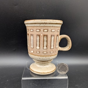 Puede incluir: Una taza de cerámica beige moteada con base de pedestal y asa. El exterior presenta un patrón repetitivo de hendiduras rectangulares verticales y pequeños cuadrados en un tono marrón más oscuro. Una moneda de veinte peniques sirve de escala.