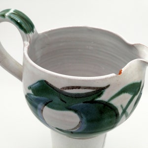 以下が含まれることがあります： 緑と青の抽象的なデザインが施された白い陶器のピッチャー。ピッチャーにはハンドルと注ぎ口があります。