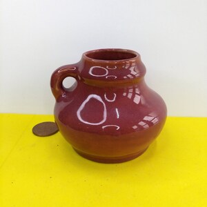 Può includere: Un piccolo boccale in ceramica color borgogna con un manico. Il boccale ha un corpo rotondo e un labbro leggermente svasato.