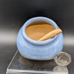 Op de afbeelding: Een kleine, ronde, blauwe keramische zoutpot met een houten lepel erin. De binnenkant is lichtbruin. De zoutpot staat op een doorzichtig plastic oppervlak. Er ligt een munt naast.
