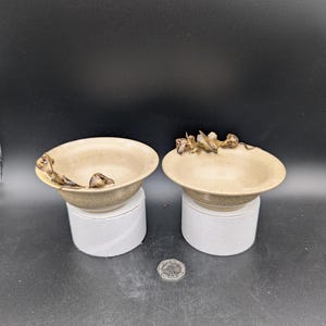 Peut inclure: Deux petits bols en céramique brun clair avec un glaçage texturé. Chaque bol a une poignée unique de forme organique en même matériau. Les bols sont posés sur des piédestaux blancs.