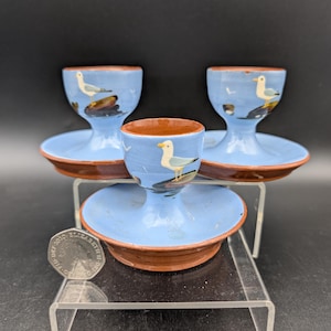 Op de afbeelding: Drie keramische eierdopjes met een lichtblauwe buitenkant en bruine binnenkant en basis. Elke beker heeft een geschilderde zeemeeuw en kustscène. De eierdopjes worden tentoongesteld op een transparante acrylstandaard.