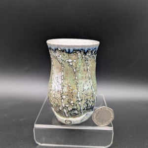 May include: A handmade ceramic cup with a unique glaze. The cup features a speckled design with shades of green, white, and blue. The rim is a light blue color. The cup is displayed on a clear stand.
