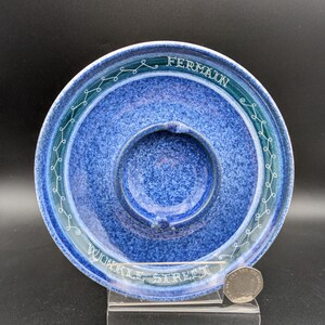 May include: A blue ceramic ashtray with a central depression and a raised outer rim. The rim features a teal band with white decorative swirls and the text "FERMAIN" and "WOKKLE STREET". A coin rests on a clear stand.