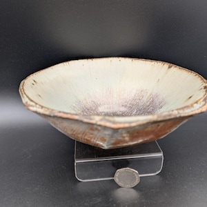 May include: A handmade ceramic bowl with a unique, irregular shape. The bowl features a light beige interior with a dark brown center and a reddish-brown exterior. The bowl is approximately 10 inches in diameter.