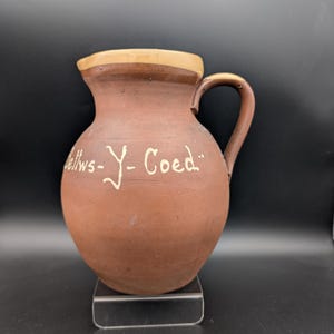 Könnte beinhalten: Brauner Keramikkrug mit rundem Körper und Henkel. Der Rand und der Henkel sind hellbraun. Die Worte "Betws-Y-Coed" sind in weißer Schrift auf der Vorderseite geschrieben. Der Krug wird auf einem transparenten Ständer präsentiert.