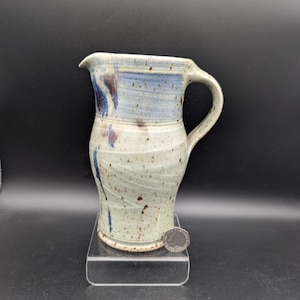 May include: A ceramic pitcher with a speckled off-white glaze, featuring blue brushwork accents and a curved handle. The pitcher is positioned next to a British twenty pence coin for scale, which has a diameter of approximately 2.14 cm.