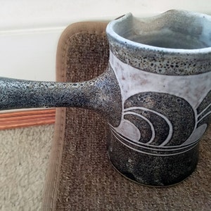 Può includere: Un'anfora in ceramica con una glassa screziata grigia e bianca. L'anfora presenta un design unico di onde nere sul lato. L'anfora ha un manico lungo.