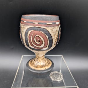 May include: A ceramic goblet with a spiral design in black and red. The goblet has a wide rim with a band of black, brown, and white. The stem and base are a lighter brown colour. The goblet is displayed on a clear acrylic stand.
