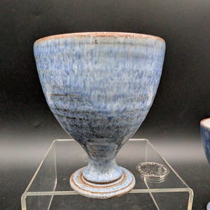 May include: Three blue ceramic goblets with a speckled glaze. The goblets have a wide bowl, a stem, and a circular base. The rims are a darker brown colour. The goblets are displayed on a clear acrylic surface.