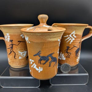 May include: A trio of ceramic items in a warm, earthy hue. The set comprises two handled mugs and a lidded jar. Each piece is decorated with black and white tribal-style illustrations of figures and animals.